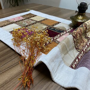 Puede incluir: Un camino de mesa blanco con un diseño de patchwork que presenta varios tonos de marrón, beige, rosa y morado. El camino de mesa tiene un borde blanco y está decorado con intrincados patrones florales. Un ramo de flores secas se coloca sobre el camino de mesa.