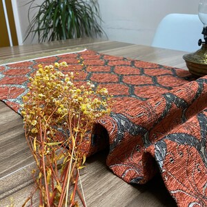 May include: A red and black patterned table runner with a fringe border. The runner is made of a textured fabric and has a geometric design. The runner is on a wooden table with a bouquet of dried flowers.