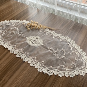 Könnte beinhalten: Ein weißer Tischläufer aus Spitze mit einem zarten Blumenmuster. Der Läufer ist aus einem durchsichtigen Stoff mit einem gewellten Rand.
