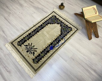 May include: A beige prayer rug with a black and green floral border and a small blue tassel. The rug has a white fringe around the edges.