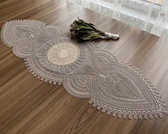 May include: A white lace table runner with a delicate floral pattern. The runner is laid out on a wooden table.