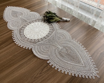 May include: White lace table runner with a delicate, intricate design featuring a large circular pattern in the center. The runner is perfect for adding a touch of elegance to any table setting.