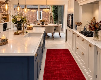 May include: A kitchen interior featuring a long, rectangular, vibrant red area rug placed on a light-colored tiled floor between a dark blue kitchen island with a white countertop and white cabinetry with gold-toned hardware.