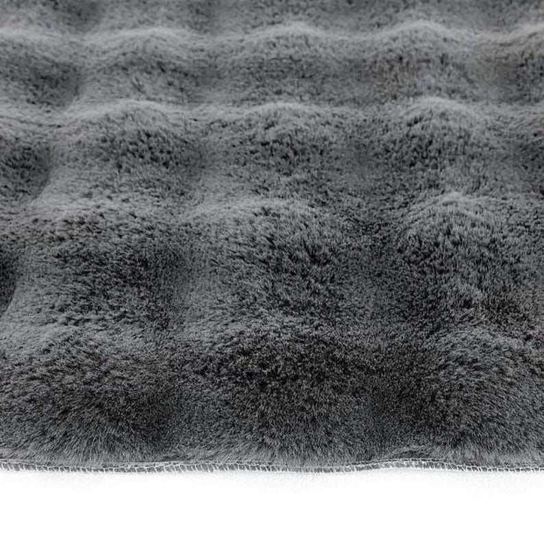 May include: Close-up of a plush, gray faux fur fabric. The fabric has a wavy texture, creating a soft, inviting appearance. The edges are neatly finished with a sewn border, suggesting a finished product like a blanket or rug.