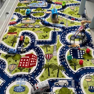 May include: A green and blue play mat with a road winding through it. The mat features various images, including cars, a helicopter, a school building, a lighthouse, and a hot air balloon. There are also numbers and math equations printed on the mat.