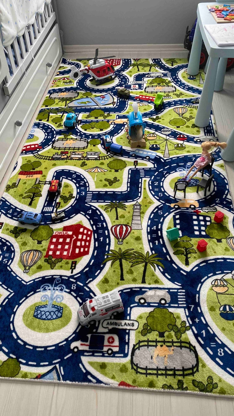 May include: A green and blue play mat with a road winding through it. The mat features various images, including cars, a helicopter, a school building, a lighthouse, and a hot air balloon. There are also numbers and math equations printed on the mat.