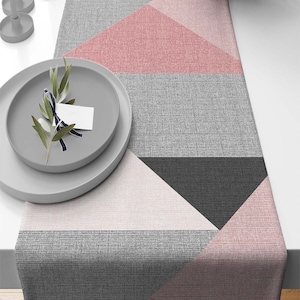 May include: A geometric table runner with a pink, gray, and black design. The runner is made of a textured fabric and has a modern, minimalist style.