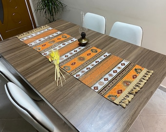 Puede incluir: Un camino de mesa marrón y naranja con un patrón geométrico y borlas. El camino de mesa está sobre una mesa de madera con sillas blancas.