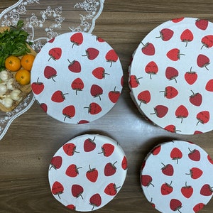 May include: Four circular fabric bowl covers featuring a white backdrop adorned with a pattern of red strawberries. The covers vary in size, designed for different bowls. Fresh produce, including lemons, garlic, and parsley, is arranged on a wooden board.