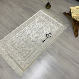Pode incluir: Um tapete de oração branco com um padrão geométrico detalhado e uma borda com franjas. Um tassel de contas de oração pretas pendura do tapete.