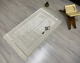 May include: A white prayer rug with a detailed geometric pattern and a fringe border. A black prayer bead tassel hangs from the rug.