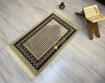 May include: A beige prayer rug with a brown and black floral pattern and a tassel. The rug has a fringed edge and a decorative arch in the center.