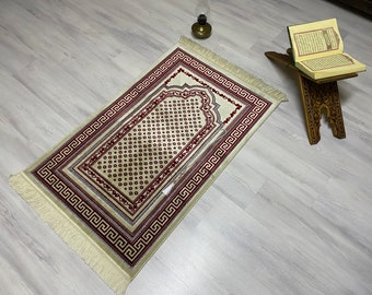 May include: A beige prayer rug with a red and white geometric pattern and fringe. The rug is laid out on a light wood floor with a wooden prayer stand and a book on top.