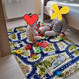 May include: Two children play on a colorful play mat with a road and city theme. The mat features numbers, a bus, an ambulance, and palm trees. The children are playing with toy cars and blocks.