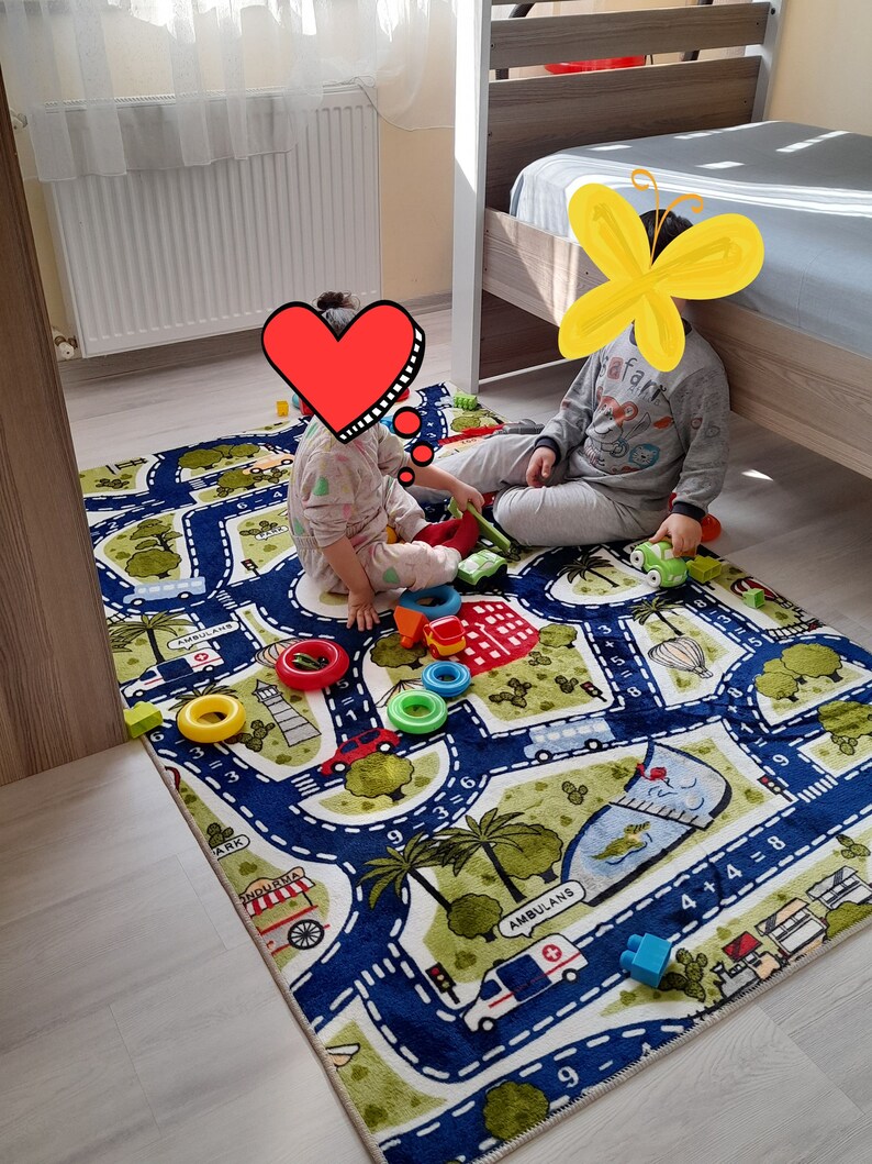 May include: Two children play on a colorful play mat with a road and city theme. The mat features numbers, a bus, an ambulance, and palm trees. The children are playing with toy cars and blocks.
