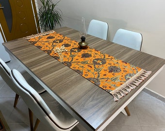 Puede incluir: Una mesa de comedor de madera marrón con sillas blancas y un camino de mesa naranja estampado con flecos.