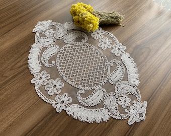 May include: White crocheted lace doily with a paisley pattern and a central circular design. The doily is on a wooden surface.
