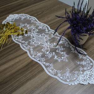 May include: A white lace table runner with a scalloped edge, featuring an intricate floral design. The runner is placed on a wooden table, alongside a small pot of purple flowers and a bundle of yellow dried flowers.