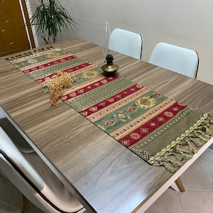 May include: A green, red, and gold patterned table runner with tassels on a wooden table. The runner has a geometric design and is placed on a wooden table with white chairs.