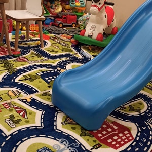 May include: A blue and green play mat with a road design and numbers. A blue slide, a red and white rocking horse, and a white toy shelf with colorful bins are in the background.