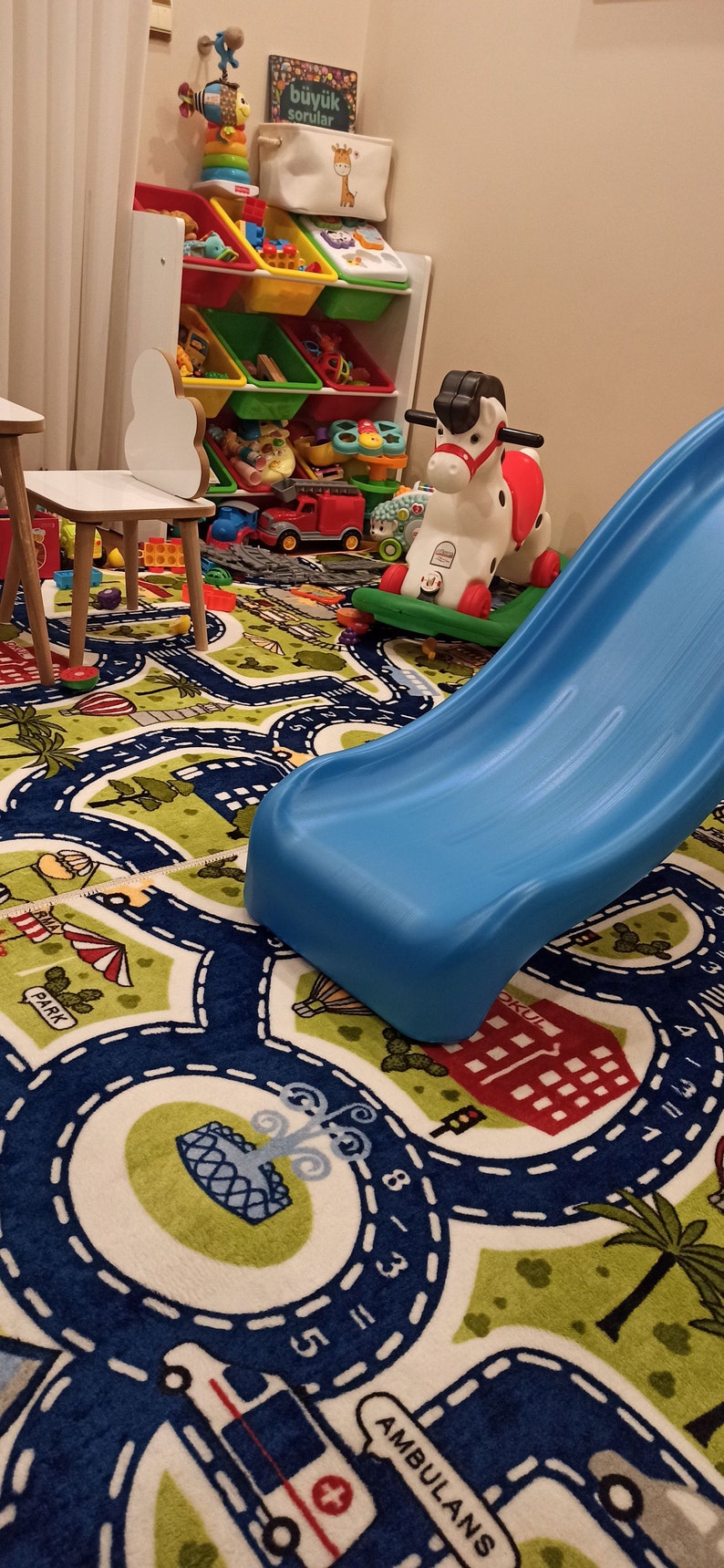 May include: A blue and green play mat with a road design and numbers. A blue slide, a red and white rocking horse, and a white toy shelf with colorful bins are in the background.
