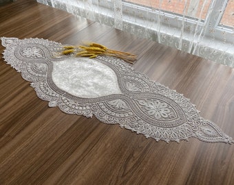 May include: White lace table runner with a delicate floral pattern. The runner is laid out on a wooden table.