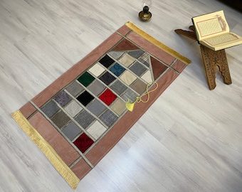 May include: A pink prayer rug with a geometric pattern of squares in various colors and textures. The rug has a gold fringe border and a yellow tassel.