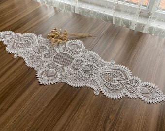 May include: White lace table runner with a delicate, intricate design. The runner features a central medallion with a repeating pattern of swirls and leaves.