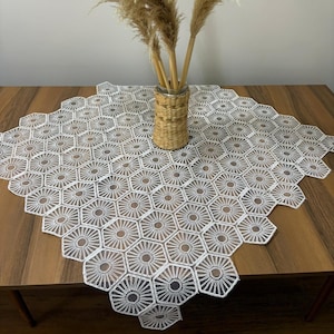 May include: A white, hexagonal lace table runner with a repeating sunburst design. The runner is on a wooden table. A woven vase with dried ornamental grass is centered on the table.