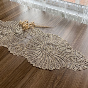 May include: A delicate, gold-colored lace table runner with intricate floral patterns. The runner is made of a sheer fabric and has a scalloped edge.