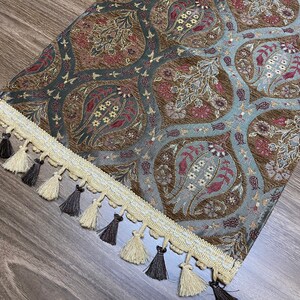 May include: A brown and blue patterned table runner with a cream and brown fringe. The runner has a floral design with a paisley-like pattern.