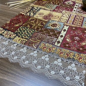 May include: A table runner with a patchwork design featuring various shades of red, brown, gold, and blue. The runner has a white lace trim.