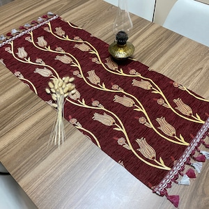 May include: A burgundy table runner with a gold floral pattern and tassel fringe. The runner is on a wooden table.