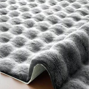 May include: A gray, plush rug with a textured, bubble-like pattern. The rug is partially rolled up, revealing a white backing and a wooden floor. The soft, fluffy material creates a cozy aesthetic.