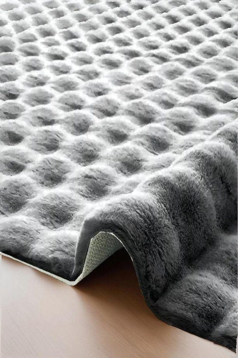 May include: A gray, plush rug with a textured, bubble-like pattern. The rug is partially rolled up, revealing a white backing and a wooden floor. The soft, fluffy material creates a cozy aesthetic.