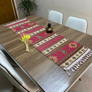 May include: A pink and cream patterned table runner with white fringe. The runner is on a wooden table with white chairs. There are dried wheat stalks on the table.
