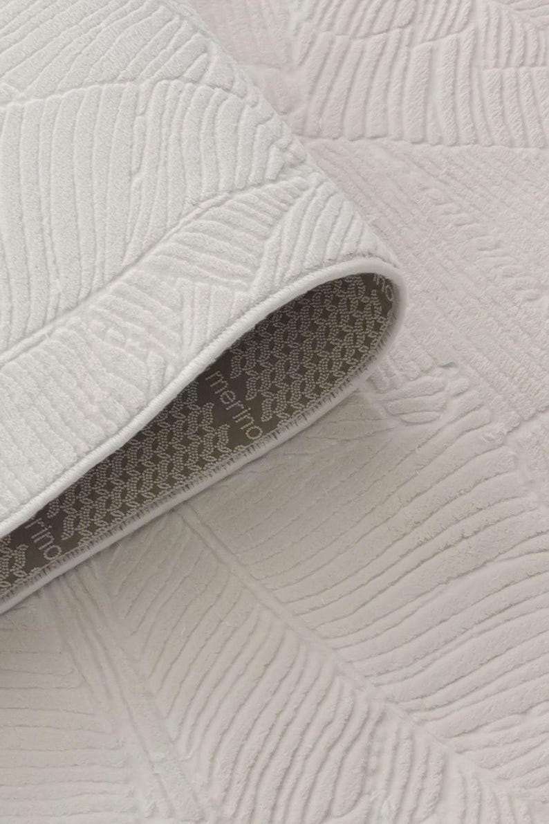 May include: Close-up of a white rug with a textured leaf pattern. The rug has a brown backing with the word "merino" printed on it.