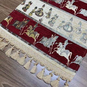 May include: A red and cream table runner with a fringe border. The runner features a repeating pattern of figures on horseback.