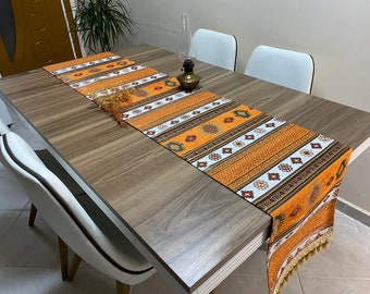 Puede incluir: Una mesa de madera marrón con un camino de mesa estampado en blanco y naranja. El camino de mesa tiene flecos en el extremo. Hay cuatro sillas blancas alrededor de la mesa.