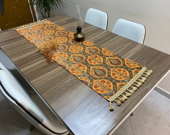 Puede incluir: Un camino de mesa marrón y naranja con un patrón floral y borlas. El camino de mesa está sobre una mesa de madera con sillas blancas.