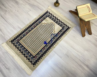 May include: A beige prayer rug with a blue and black floral pattern and a blue tassel. The rug has a fringe border and is laid out on a light wood floor.