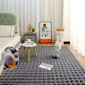 May include: A large, rectangular gray faux fur rug with a textured, tufted pattern. The rug is placed on a wooden floor in a bedroom setting. A white bed, side table, and artwork are visible in the background.