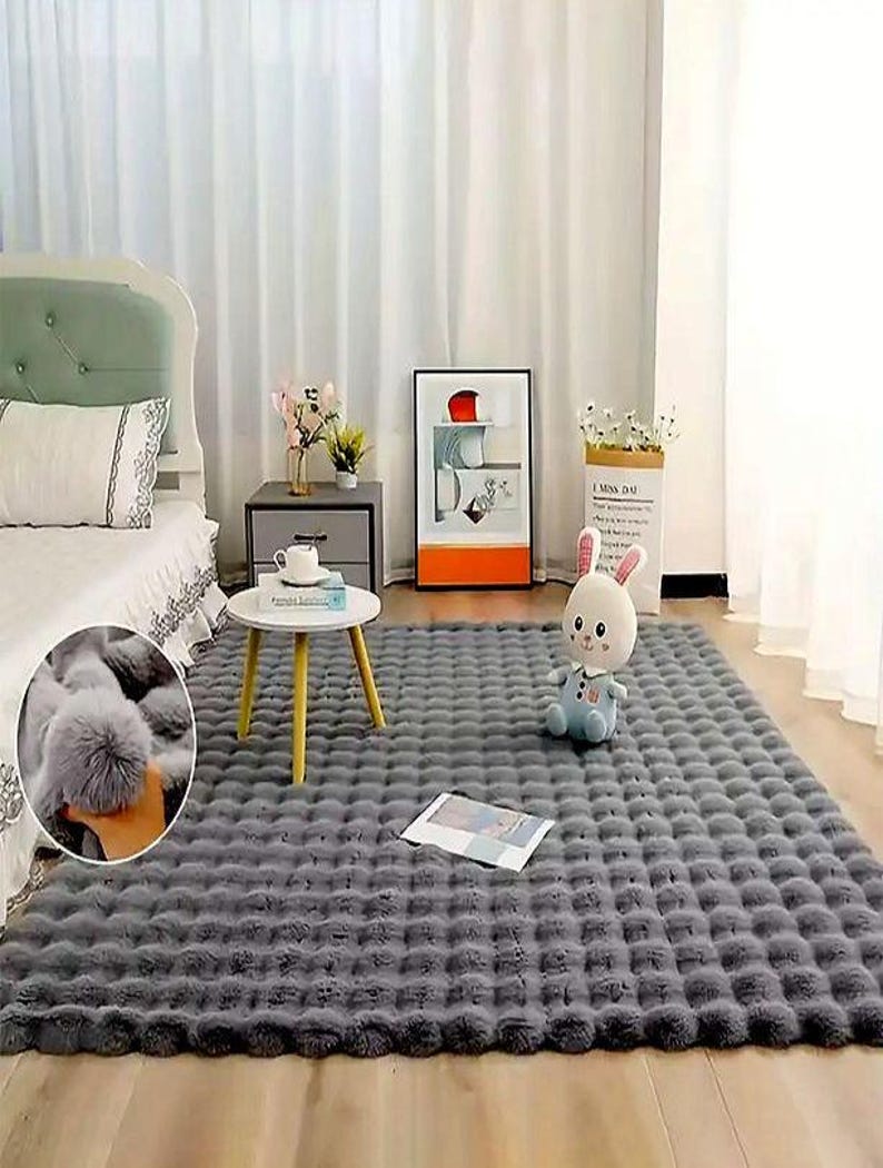 May include: A large, rectangular gray faux fur rug with a textured, tufted pattern. The rug is placed on a wooden floor in a bedroom setting. A white bed, side table, and artwork are visible in the background.