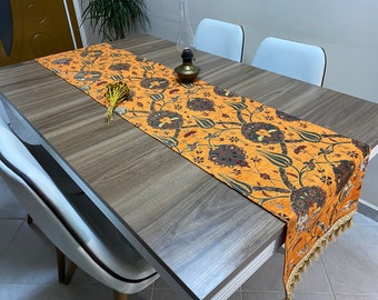 Puede incluir: Un camino de mesa largo y naranja con un diseño turco tradicional que presenta intrincados patrones florales en tonos de marrón, rojo y verde. El camino de mesa tiene un borde con flecos y está colocado sobre una mesa de madera.
