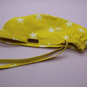 Op de afbeelding: Gele scrub-muts met witte sterren en een bruin label met de tekst "SCRUB LIFE". De muts heeft een knoop en banden.