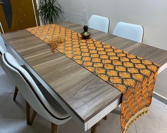 May include: Orange and black patterned table runner with gold fringe on a wooden table with white chairs.