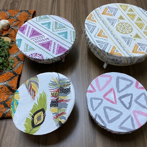 May include: Four reusable food covers with different patterns. The covers are made of fabric and have a white background. The patterns include geometric shapes, feathers, and abstract designs. The covers are a great way to keep food fresh and prevent spills.