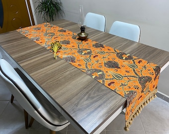 Puede incluir: Un camino de mesa largo de color naranja con un diseño turco tradicional en tonos marrones, verdes y rojos. El camino de mesa tiene un borde de flecos y está colocado sobre una mesa de madera.