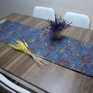 May include: A blue table runner with a floral pattern and fringe edges. The runner is on a wooden table with a vase of lavender flowers and wheat stalks.