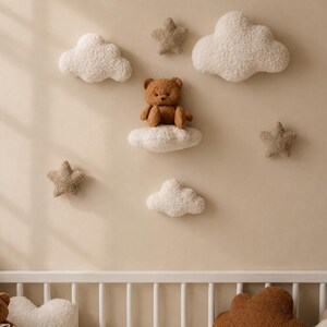 Op de afbeelding: Een muurdecoratie voor de kinderkamer met een bruine teddybeer die op een wit wolk-vormig kussen zit, omringd door zachte wolken en stervormige kussens. Een banner van driehoekige vlaggen hangt erboven. Het kleurenpalet is neutraal met beige en bruine tinten.